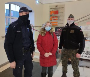 Zdjęcie przedstawia trzy osoby stojące w wjściu galerii handlowej. Pierwsza osoba od lewej to policjant ubrany w granatowy mundur i granatową kurtkę. Na głowie ma granatową czapkę z daszkiem i białym napisem POLICJA umieszczonym po prawej stronie. Na twarzy ma maseczkę w kolorze czarnym W środku stoi kobieta w okularach i błękitnej maseczce na twarzy. Włosy koloru blond sięgające ramion. Ubrana w czerwoną kurtkę do połowy ud z puchowym kołnierzem koloru brązowo-żółtego i spodnie koloru czarnego. Kolejna osoba stojąca po prawej stronie to żołnierz ubrany w czarną, polarową bluzę. Na lewym ramieniu widoczna naszywka z flagą Polski. Spodnie moro w kolorze zielono-brązowym. Na głowie ma furażerkę moro z widocznym orzełkiem z przodu, a na twarzy maseczkę. W tle po lewej stronie widoczne jest okno. Po środku nad schodami zawieszony jest plan galerii handlowej. Rozkład pomieszczeń jest w kolorach pomarańczowym i czerwonym. Po prawej stronie widoczna metalowa poręcz. Ściany są w kolorze beżowym.