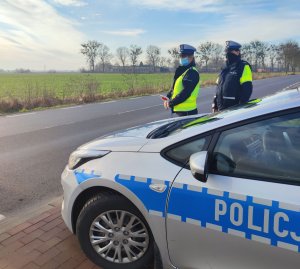 Na pierwszym planie zdjęcia widoczny jest lewy bok radiowozu stojącego na poboczu, przodem do drogi asfaltowej. Radiowóz koloru srebrnego z biegnącym wzdłuż niebieskim paskiem, na którym widnieje biały napis POLICJA. Za radiowozem stoją dwie policjantki z ruchu drogowego. Częściowo schowane za pojazd, ubrane w granatowe mundury i białe czapki. Jedna z policjantek trzyma w dłoni biało-czerwoną tarczę do zatrzymywania pojazdów. W tle widoczna droga asfaltowa z poboczem, zielone pole uprawne. W dalszej odległości widoczne zabudowania oraz pasmo drzew.