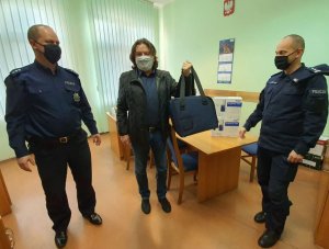 Zdjęcie przedstawia trzech mężczyzn w pomieszczeniu biurowym. Dwóch z nich to policjanci w granatowych mundurach i maseczkach na twarzach. Po środku stoi mężczyzna ubrany w czarną kurtkę i niebieskie spodnie. Na twarzy ma maseczkę. W ręku trzyma przenośną torbę z ufundowanym analizatorem.