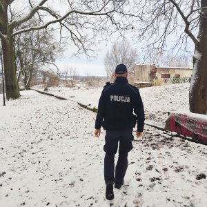 Zdjęcie przedstawia umundurowanego Policjanta idącego po śniegu w kierunku opuszczonego budynku. Budynek jest niski, zniszczony i otoczony drzewami.