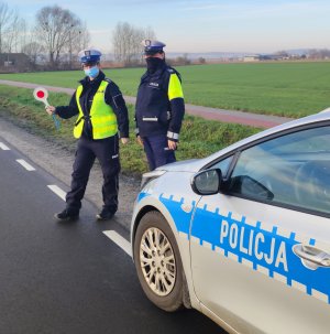 Zdjęcie przedstawia dwie funkcjonariuszki ruchu drogowego, ubrane w granatowe mundury, białe czapki i elementy odblaskowe. Jedna z policjantek trzyma w dłoni tarczę do zatrzymywania pojazdów. Policjantki stoją przed radiowozem, przy drodze asfaltowej. W tle widoczna ścieżka rowerowa, drzewa, domostwa oraz pole.