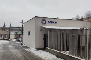 Zdjęcia poglądowe przedstawiają biały budynek z niebieskim napisem POLICJA. Nad bramką wejściową znajduje się zadaszenie. Widoczne elementy ogrodzenia.