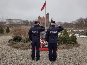 dwaj komendanci stoją tyłem do fotografa i oddają honory przed pomnikiem poświęconym pamięci Żołnierzy Wyklętych na Skwerze im. Konfederatów Obrony Wiary i Ojczyzny przy ul. Długiej w Marezie i powiewającą na maszcie flagę, w tle widać kwidzyński zamek