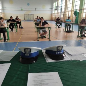 Zdjęcie podglądowe przedstawia dwie czapki funkcjonariuszy Wydziału Ruchu Drogowego, leżące na stole wraz z testem egzaminacyjnym. W tle widać dzieci siedzące przy stolikach i rozwiązujące test. Na drugim zdjęciu widać plac, z wyznaczonymi torami jazdy, a także dziecko stojące przy rowerze w kasku na głowie i ubranej kamizelce odblaskowej oraz policjantkę ruchu  drogowego, która egzaminuje uczestników konkursu.
Na trzecim zdjęciu widoczny uczestnik konkursu poruszający się po placu rowerem oraz policjantka ruchu drogowego. Na czwartym zdjęciu widoczni dwaj uczestnicy konkursu poruszający się po torze przeszkód. Obok stoi policjant ruchu drogowego. Na piątym zdjęciu widoczny jest plac egzaminacyjny, dziecko jadące na rowerze. Przy placu oraz na placu stoją policjnci ruchu drogowego.