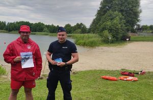 Zdjęcie poglądowe przedstawia ratownika wodnego oraz policjanta. Obaj stoją na plaży przy jeziorze Kamień w Gardei. W rękach trzymają plakaty oraz ulotki dotyczące akcji &quot;Kręci mnie bezpieczeństwo nad wodą&quot;