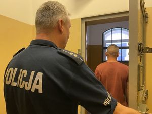 Zdjęcie przedstawia stojącego tyłem policjanta trzymającego otwarte drzwi do celi. W progu celi stoi odwrócony tyłem mężczyzna ubrany w koszulkę w kolorze pomarańczowym, który jest sprawcą włamania.