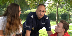 na zdjęciu widać opartego rękoma o tył ławki w parku policjanta po lewej stronie funkcjonariusza siedzi na ławce młody chłopak  w czerwonej bluzie i mu się przygląda po prawej stronie policjanta siedzi na oparciu ławki młoda dziewczyna trzyma telefon w ręku i również przygląda się funkcjonariuszowi. W tle zdjęcia widać zieloną roślinność w parku.