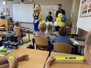 zdjęcie zrobione w klasie w której siedzą w ławkach uczniowie a stoją policjanci i osoba przebrana za żółtą maskotkę mrówki