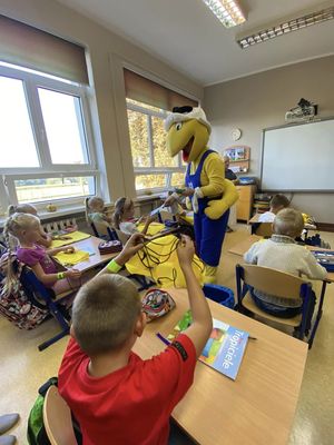 zdjęcie przedstawia osobę przebraną za dużą żółtą maskotkę mrówki która rozdaje dzieciom w klasie odblaski