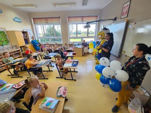 zdjęcie zrobione w klasie w której siedzą w ławkach uczniowie a stoją policjanci i osoba przebrana za żółtą maskotkę mrówki