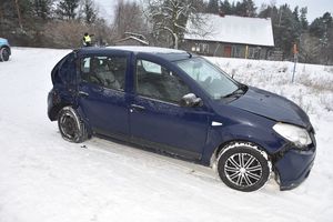 na zaśnieżonej drodze stoi niebieski samochód z uszkodzeniami pokolizyjnymi w tle widać policjanta budynek i las