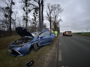 na zdjęciu widać rozbity samochód z uszkodzoną lewą stroną stojący na poboczu obok niego widać strażaków a w tle drugi samochód który brał udział w tym samym zdarzeniu na poboczu i w głębi planu widać drzewa