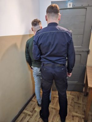 Zdjęcie, na którym policjant wprowadza zatrzymanego mężczyznę do pomieszczenia dla osób zatrzymanych.