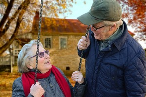 na zdjęciu para seniorów starsza kobieta siedzi na huśtawce a obok niej stoi starszy pan i jedną ręką trzyma za sznurek huśtawki