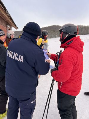 na zdjęciu widać umundurowanego policjanta stojącego obok narciarza któremu wręcza ulotkę dotyczącą zasad na stoku, w oddali widać stok narciarski pokryty śniegiem