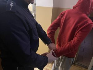 na zdjęciu widać umundurowanego policjanta, który przed celą dla osób zatrzymanych zakłada mężczyźnie ubranemu w czerwoną bluzę i spodnie dresowe kajdanki na ręce trzymane z tyłu