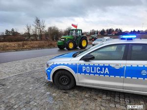 na zdjęciu widać zaparkowany na poboczu radiowóz oraz jadący ulicą ciągnik, który ma na dachu flagę biało czerwoną a za nim samochód osobowy