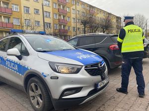 policjant przechodzący obok radiowozu w stronę samochodu który jest obok niego zaparkowany na parkingu a jechał nim nietrzeźwy kierujący