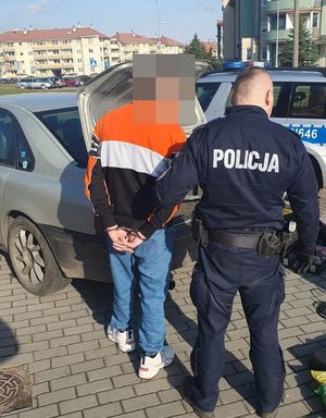 na zdjęciu widać oprócz zatrzymanego samochodu stojącego obok policjanta mężczyznę który posiadał narkotyki ma na ręce założone kajdanki a przed nimi stoi radiowóz policyjny