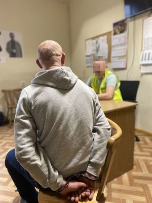 na zdjęciu widać mężczyznę który ma założone na ręce kajdanki i siedzi na krześle a przed nim za biurkiem siedzi policjant