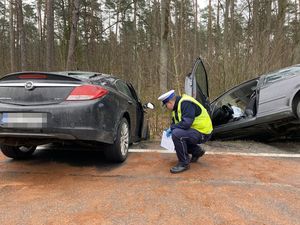 pomiędzy dwoma samochodami które czołowo się zderzyły i wiszą przodem na rowem kuca umundurowany policjant i sprawdza opony auta