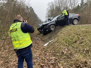 pomiędzy dwoma samochodami wiszącymi na tylnych kołach nad rowem stoi policjant w kamizelce odblaskowej i robi zdjęcia uszkodzeń