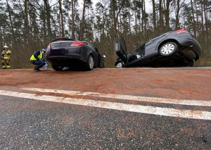 na zdjęciu widać dwa auta które się zderzyły i tylnymi kołami stoją na jezdni a przednia część oby samochodów wisi nad rowem obok jednego auta po prawej stronie kuca policjant