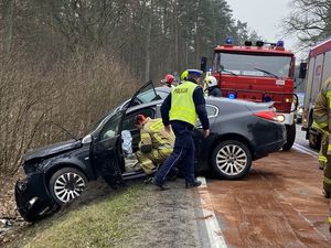 na zdjęciu widać rozbity samochód obok niego kucający strażak zagląda do środka tuż obok przechodzi policjant w tle widać wóz strażacki a na ulicy rozsypany piasek absorbujący wycieki z aut