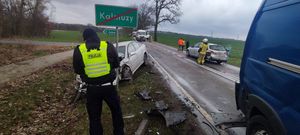 na pierwszym planie widać tył samochodu dostawczego następnie po lewej stronie opisującego uszkodzenia rozbitego samochodu osobowego znajdującego się przed nim i znak drogowy z napisem kalmuzy w tle widać inne auta i strażaków