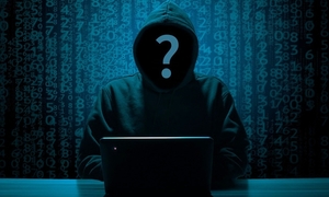 zdjęcie w ciemnej kolorystce przed komputerem siedzi mężczyzna w bluzie z kapturem w miejscu twarzy jest znak zapytania