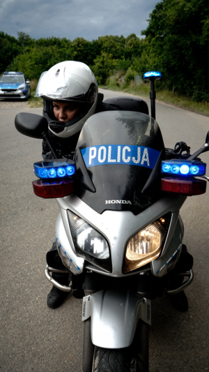 na zdjęciu widać policjantkę siedzącą w białym kasku na policyjnym motocyklu zaparkowanym na asfalcie w tle widać radiowóz i drzewa