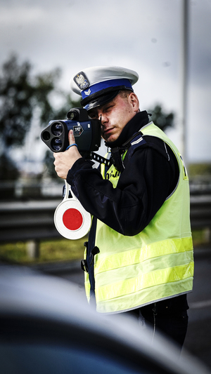 na zdjęciu widać umundurowanego policjanta który stoi z przyłożonym do oka miernikiem służącym do pomiaru prędkości samochodów w ręku trzyma też luźno zwisający na sznurku lizak policyjny służący do zatrzymywania pojazdów