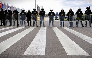 na pierwszym planie widać jezdnię na której namalowane są białe pasy dalej stoją w rzędzie policjanci którzy opierają ręce na tarczy na głowach mają białe kaski są ubrani w mundur