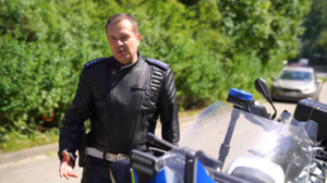 na zdjęciu widać stojącego po lewej stronie motocykla policjanta ubranego w skórzany kombinezon w tle widać ulicę i krzaki