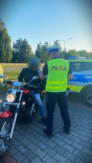 na zdjęciu widać umundurowanego policjanta stojącego tyłem na parkingu przy radiowozie a przed nim siedzącego w kasku z podniesioną przyłbicą na motocyklu mężczyznę który dmucha w alkomat