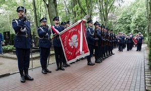 zdjęcie zrobione na cmentarnej alejce na której są poustawiani w szeregu i dwuszeregu umundurowani policjanci jeden z nich trzyma sztandar w ręku dalej widać policjantów z orkiestry dętej w tle drzewa i nagrobki