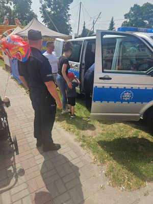 na zdjęciu widać stojącego policjanta obok radiowozu do którego wchodzi dziecko w towarzystwie swoich rodziców w tle kolorowe namioty wystawione na czas trwającego festynu