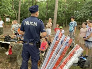 policjant prowadzi lekcję historii wśród dzieci siedzących na kocach w lesie widać stolik na którym są historyczne umundurowania i broń z czasów wojny oraz wiele innych ciekawych rzeczy na pierwszym planie widać elementy odblaskowe które otrzymają na zakończenie spotkania dzieci