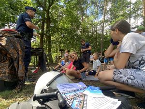 policjant prowadzi lekcję historii wśród dzieci siedzących na kocach w lesie widać stolik na którym są historyczne umundurowania i broń z czasów wojny oraz wiele innych ciekawych rzeczy