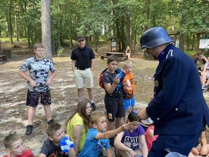 policjant prowadzi lekcję historii wśród dzieci siedzących na kocach w lesie widać stolik na którym są historyczne umundurowania i broń z czasów wojny oraz wiele innych ciekawych rzeczy