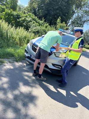 zdjęcie przedstawia policjantkę obok radiowozu która zatrzymała rowerzystę do kontroli drogowej mężczyzna ten jest nachylony nad maską radiowozu i podpisuje mandat