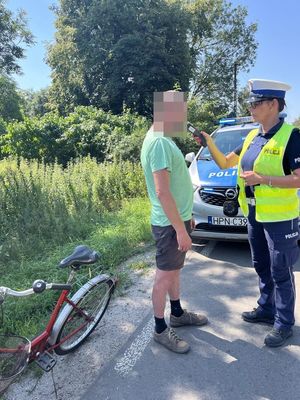 zdjęcie przedstawia policjantkę która trzyma w ręku alkomat i podaje go mężczyźnie żeby dmuchnął w ustnik w celu sprawdzenia stanu trzeźwości obok mężczyzny stoi rower w tle widać radiowóz