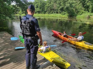 na zdjęciu widać stojącego na brzegu rzeki tyłem do fotografa policjanta oraz płynących dwoma kajakami ludzi w tle widać lasy