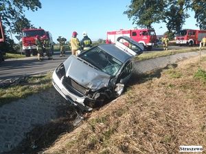 na zdjęciu widać samochód kolor srebrnego który jest przewrócony na bok w rowie melioracyjnym w tle stoją trzy wozy strażackie i strażacy