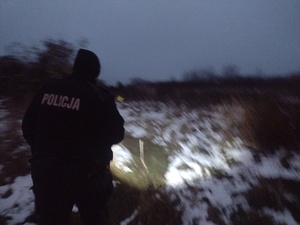 na zdjęciu widać umundurowanego policjanta który idzie z latarką przez łąkę na trawie widać śnieg