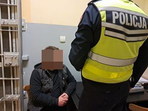 na zdjęciu widać mężczyznę siedzącego na krześle trzyma on ręce razem oparty łokciem o biurko za nim są okratowane drzwi do celi a przed mężczyzną stoi umundurowany policjant
