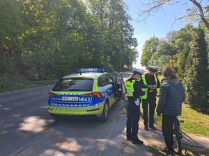 na pierwszym planie zdjęcia widać mężczyznę stojącego obok roweru tuż obok dwie policjantki stojące przy radiowozie który ma otwarte drzwi od strony pasażera funkcjonariuszki legitymują mężczyznę w tle widać ulicę i duże drzewa