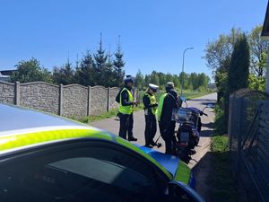 na pierwszym planie widać fragment policyjnego radiowozu a dalej funkcjonariuszki które legitymują mężczyznę zatrzymanego do kontroli drogowej który jechał skuterem