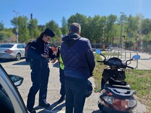 na zdjęciu widać policjantki z czego jedna sprawdza dowód osobisty zatrzymanego do kontroli drogowej mężczyzny który jechał skuterem w tle widać zaparkowane na parkingu samochody
