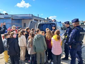 na zdjęciu widać troje policjantów którzy pokazują zaparkowany na policyjnym parkingu radiowóz dzieciom które przyjechały na wycieczkę by zapoznać się z tajnikami pracy policjantów